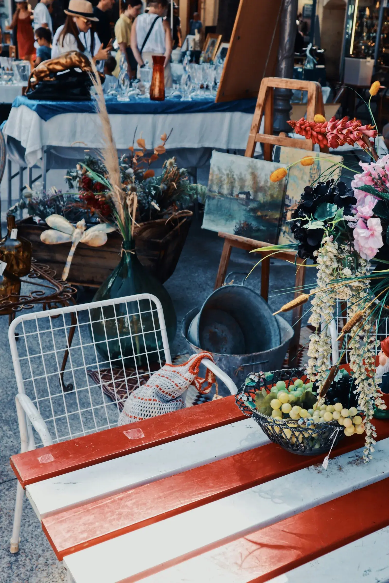 A brocante flea-market stall with dried flower arrangements, a vintage glass demijohn, antique paintings, and a wire basket of grapes on a striped bench
